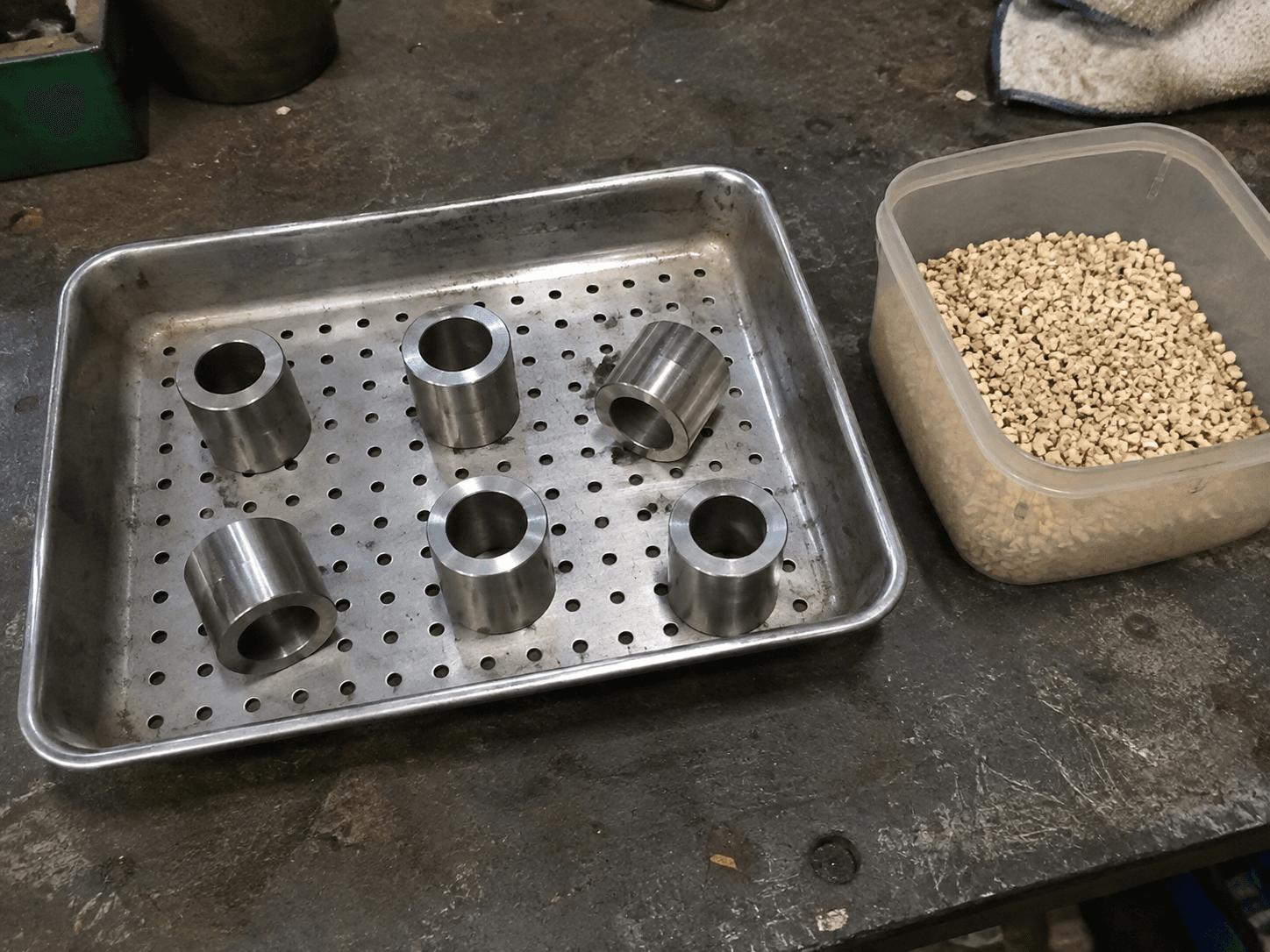 Generic metal sleeves in a perforated tray beside drying granules on a workshop bench.
