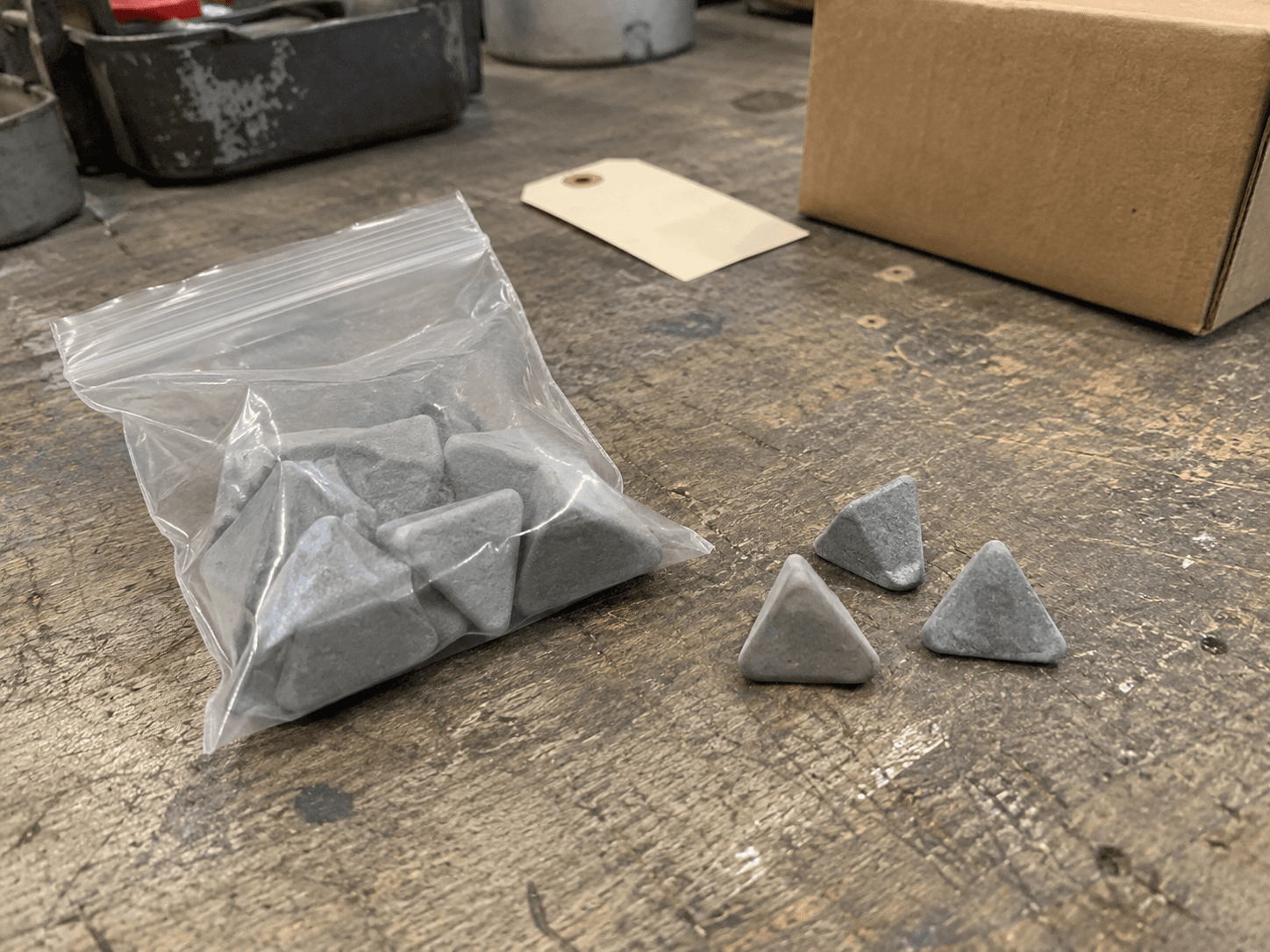 AI illustration of gray triangular media in a clear bag and three loose pieces on a worn workshop bench beside a blank tag and box.