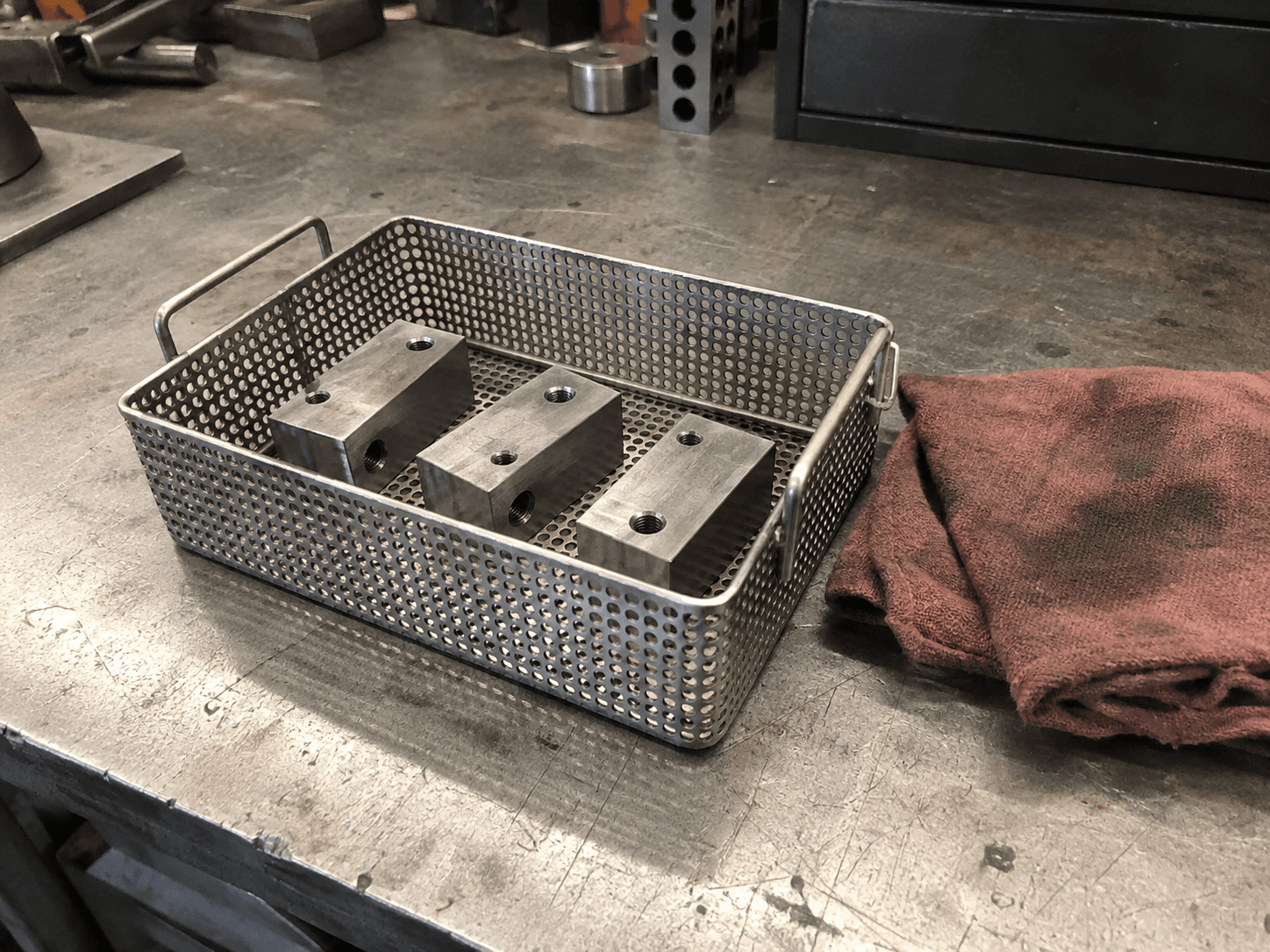Generic machined blocks in a perforated parts basket beside a shop cloth.