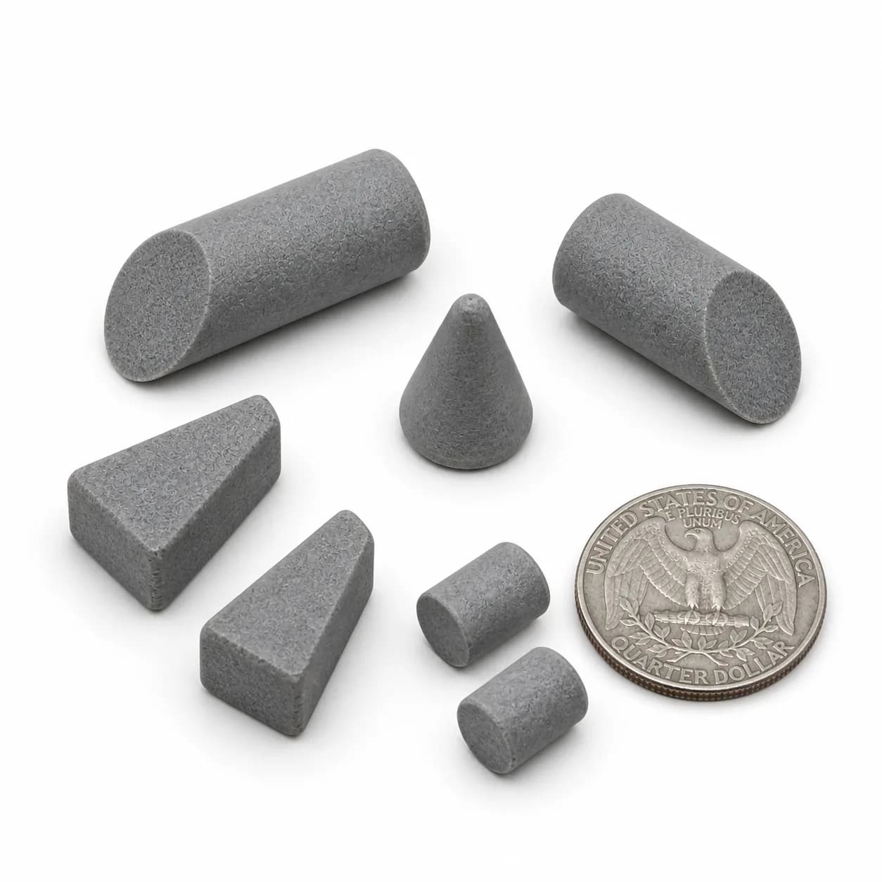 Five gray finishing media pieces in triangle, cylinder, and cone shapes arranged beside a US quarter