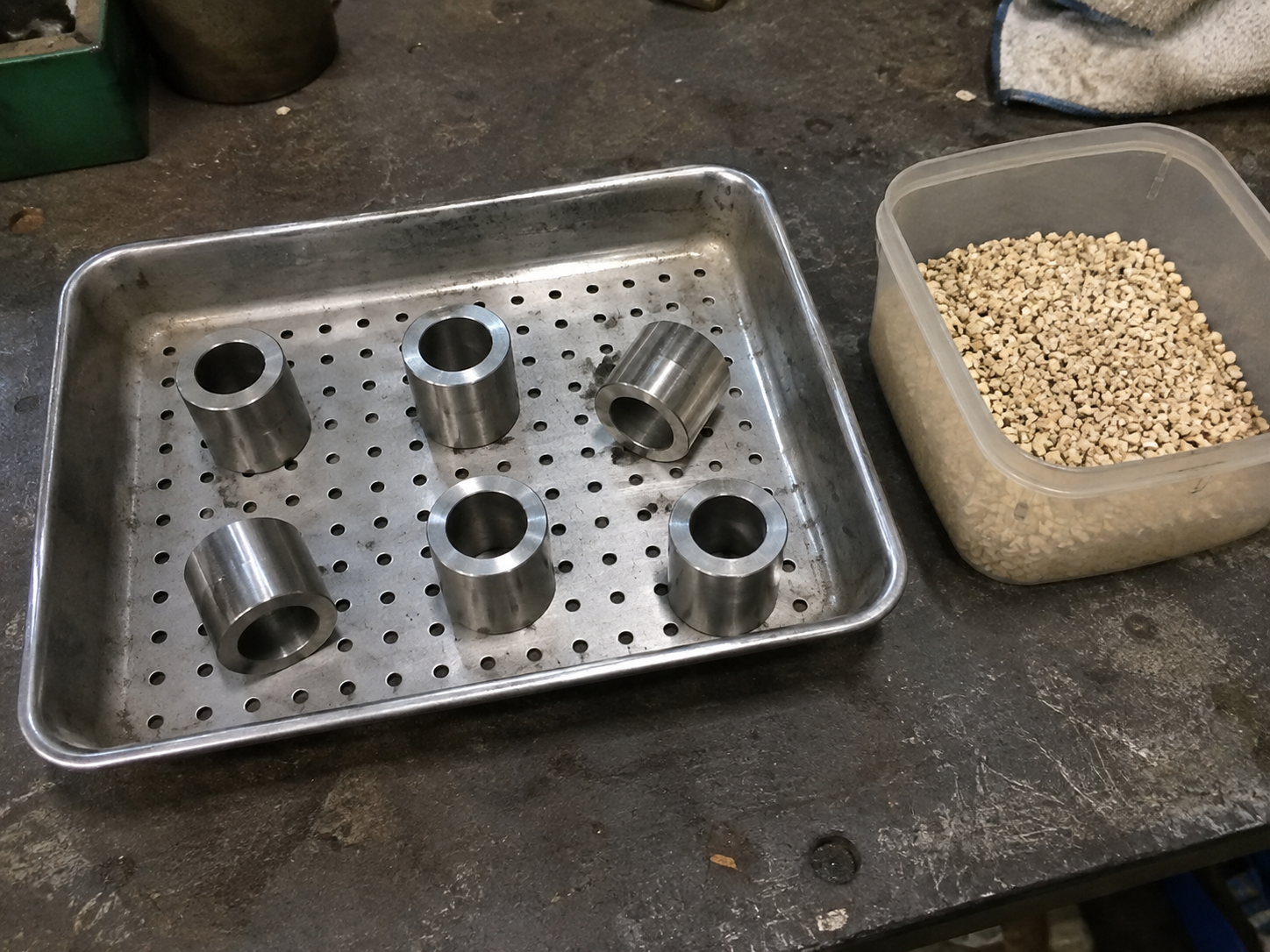 Generic metal sleeves in a perforated tray beside drying granules on a workshop bench.